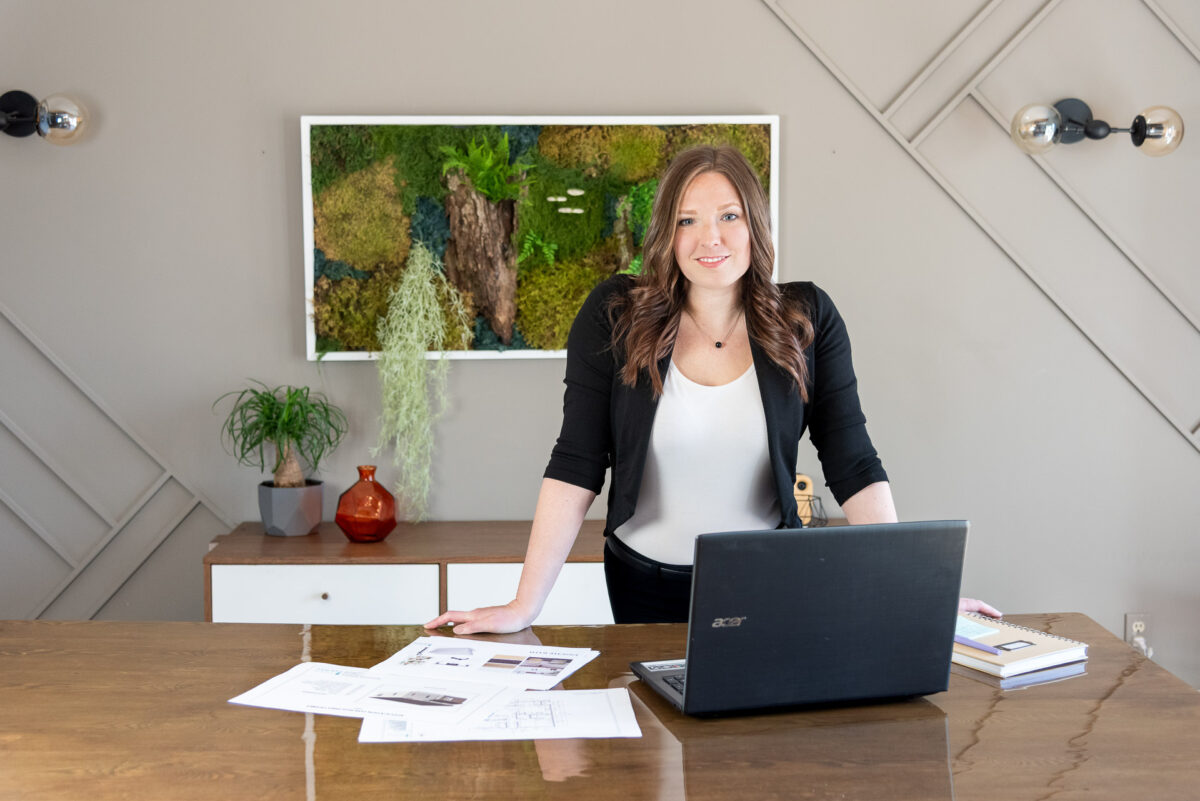 woman with brown hair wearing a black and white top leaning on a table. Behind her is a living wall of mossy plants. On the table is an open laptop and printed schematics