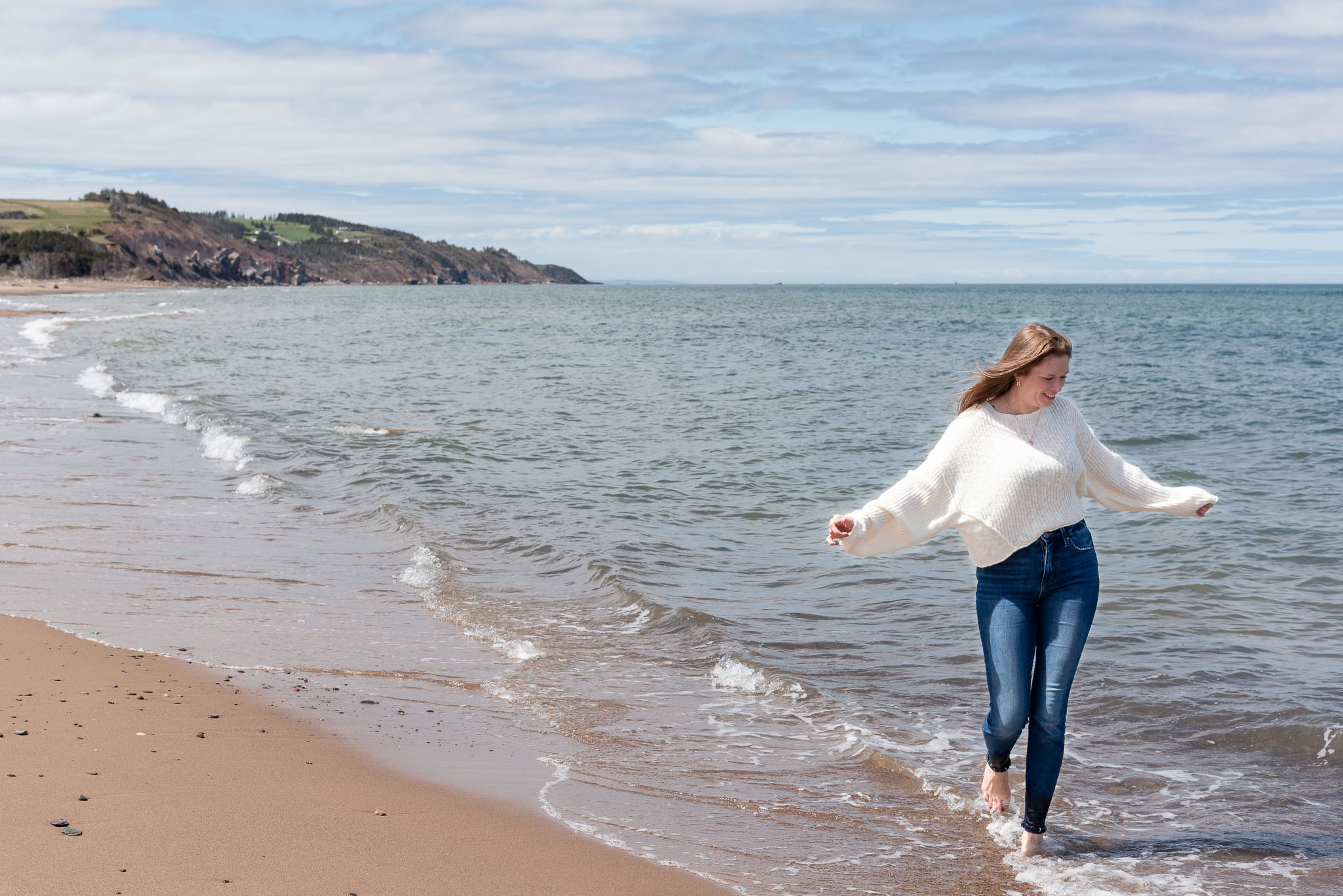 female birth coach walking in the waves in Cape Breton, wearing jeans and a white sweater, during a Brand Visibility photoshoot with Dandelion Digital
