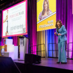 Keynote speaker Barb Stegemann on stage. Behind her are colourful, bright wall drapes and her photo on screen