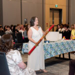 a women in the centre of a crowded room, wearing a white linen dressing, holding a rain stick
