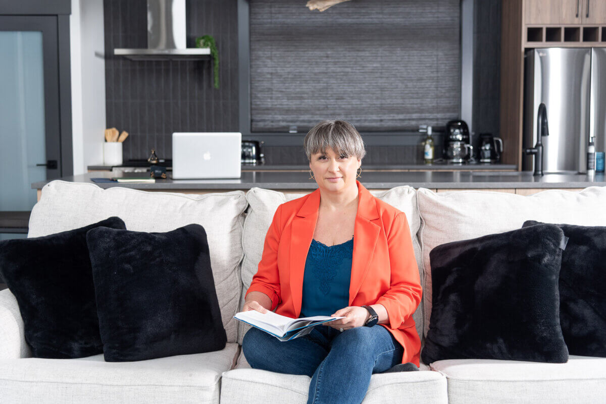 woman in an orange jacket and jeans, taking notes on a sofa with a counter in the background with an open macbook