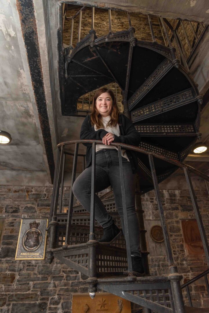 dark haired, young woman wearing grey branded sweater, on spiral staircase