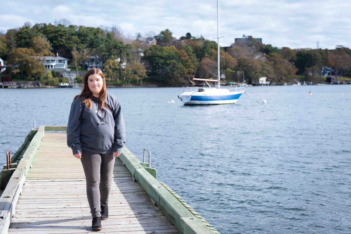 dark haired, young woman wearing grey branded sweater, walking on dock with sailboat on lake in background