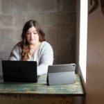 dark haired, young woman wearing grey sweater sitting at desk working with 2 iPads