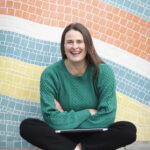a women sitting crossed legged against a colourful tile wall with arms crossed on laptop on her lap