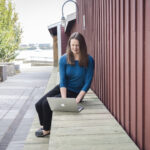a women with long brown hair, wearing a royal blue top and black pants, sitting on a bench typing on her laptop. The Halifax harbour is in the background