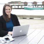 a women with long brown hair, wearing a royal blue top, black jacket and black pants, sitting at an outdoor table with her laptop. Behing her is the Halifax harbour with a small baot tied to a pier