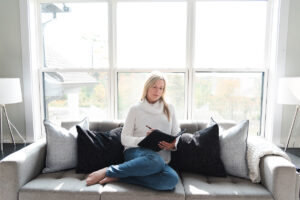 blond woman in a white sweater and blue jeans, with her legs curled under her on the couch as she writes in a black journal