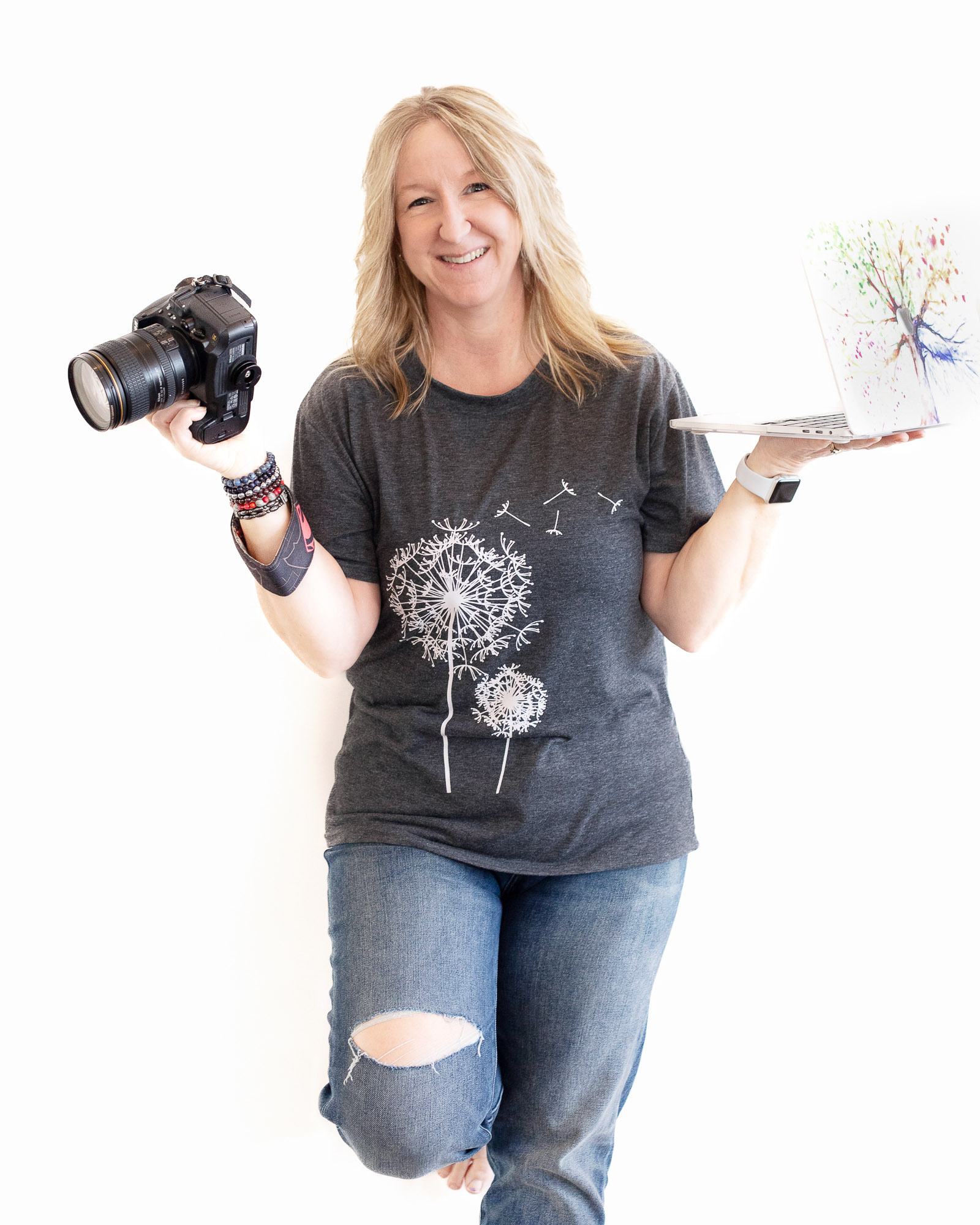 Allison Smith is a blonde photographer in a black t-shirt with a white dandelion holding a camera in one hand and balancing a laptop in the other.