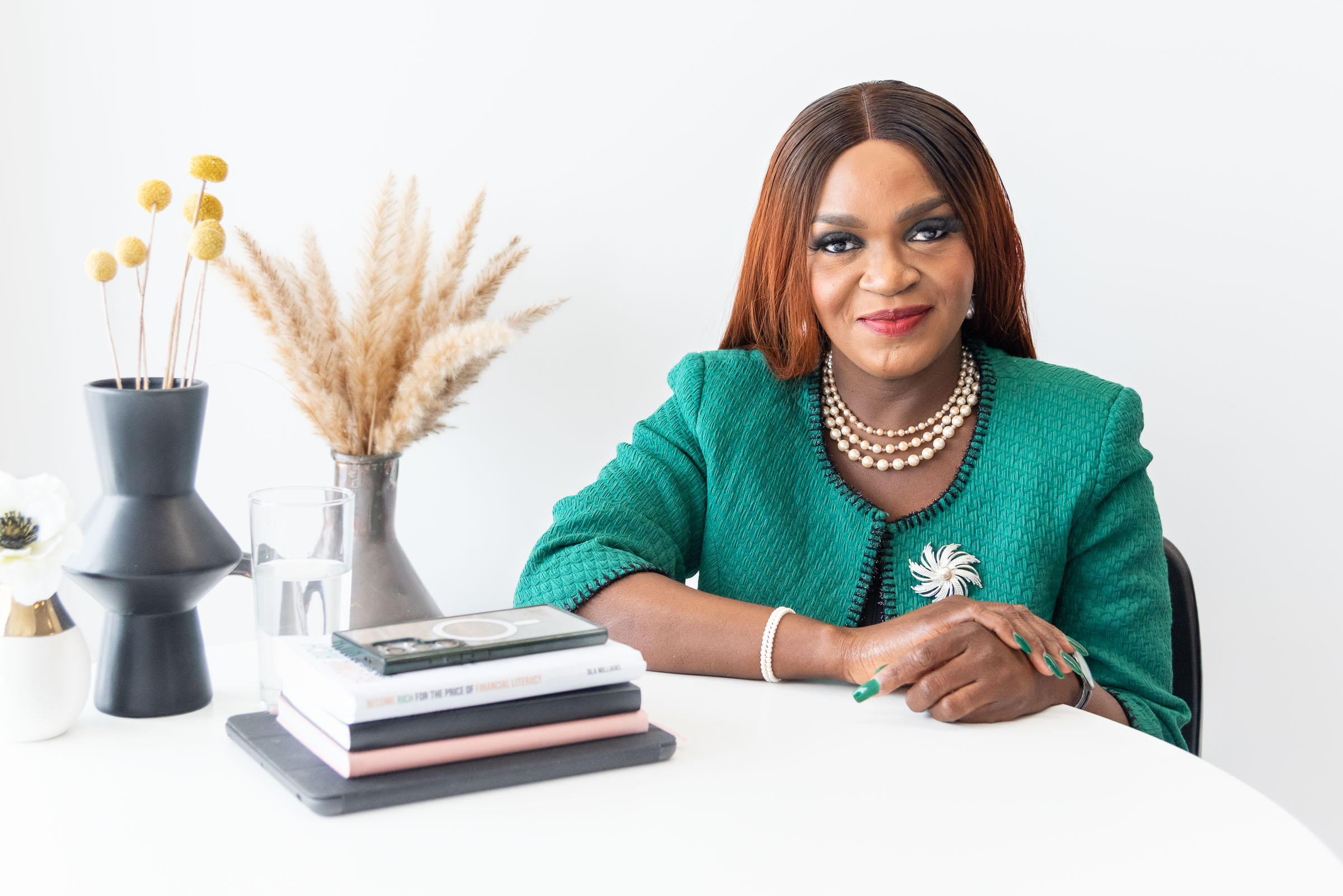 profile photo of a black women in a green blazer sitting at a desk with books and decorative plants on the table