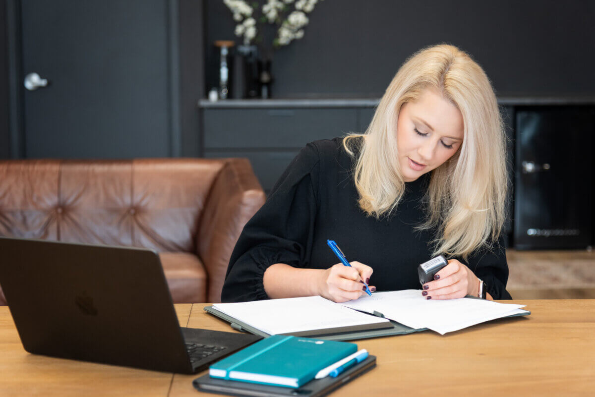 blonde female lawyer in a balck dress signing documents at a wood desk with a brown leather sofa behind her