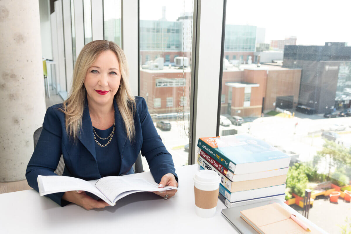 blond woman in dark suit flipping through a book at a desk in the library along a wall of windows