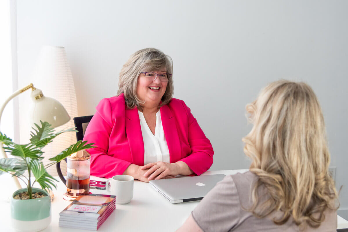 woman in a hot pink jacket sitting at a desk with a macbook, plant, lamp and stack of her published across. Across from her you see the back of her client