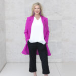 blond women in hot pink duster jacket, white blouse & black pants in a standing pose in front of marble wall for photography subscription shoot
