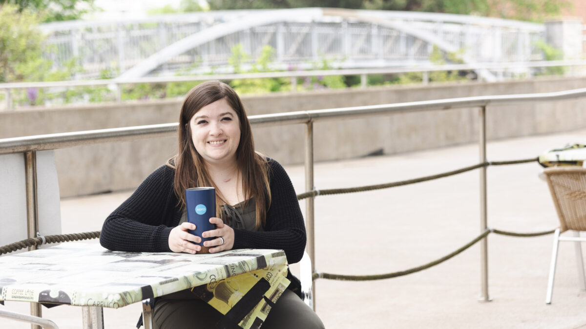 dark haired, young woman sitting at a coffee shop's outdoor patio with a walking bridge in the background