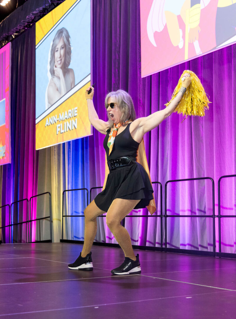 a women on stage in a short black dress with yellow pompoms in her raised arms during movement break at Wonder Women conference, Halifax Convention Centre