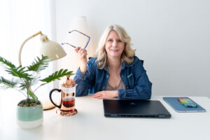 blond women in a denim shirt holding her glasses in her right hand while sitting at a white table. On the table is a closed laptop, coffee press, lamp and plant. 