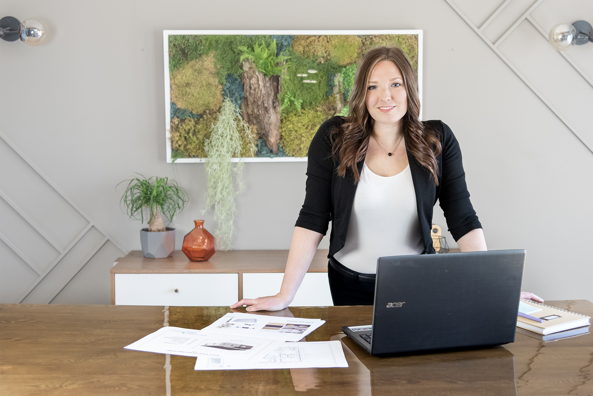 Dandelion Digital Mini branding session at Creation Lounge, Hammonds Plains, NS. Dark haired woman wearing a white top and black blazer leaning on a table. Behind her is a living wall of mossy plants. On the table is an open laptop and printed schematics