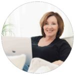 woman with short brown hair, leaning back on a sofa, with a laptop on her lap, smiling at the camera