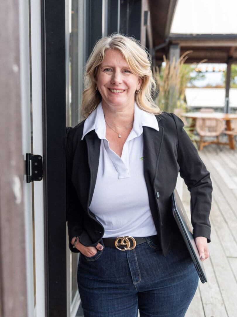 blond woman in dark suite leaning on wall, holding laptop on outdoor patio