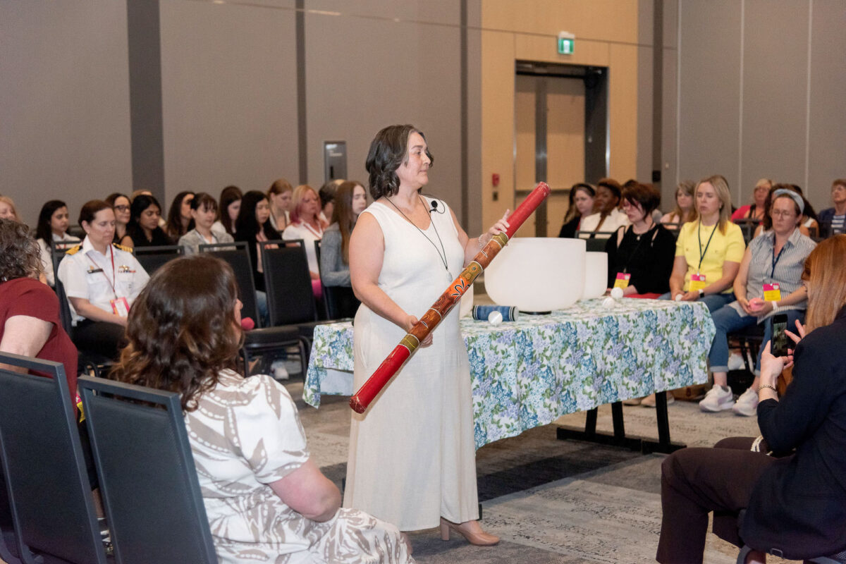a women in the centre of a crowded room, wearing a white linen dressing, holding a rain stick during Halifax Chamber of Commerce Wonder Women Conference meditation session
