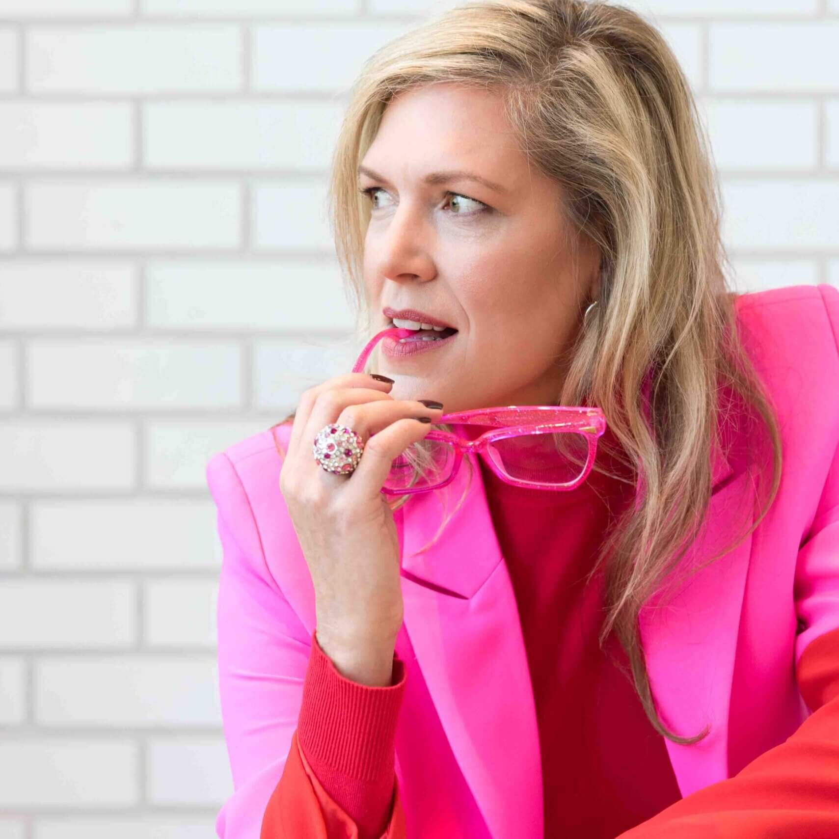 headshot of a blond women looking off to the side, chewing tip of hot pink fashion glasses. She is wearing a pink jacket with red turtle neck