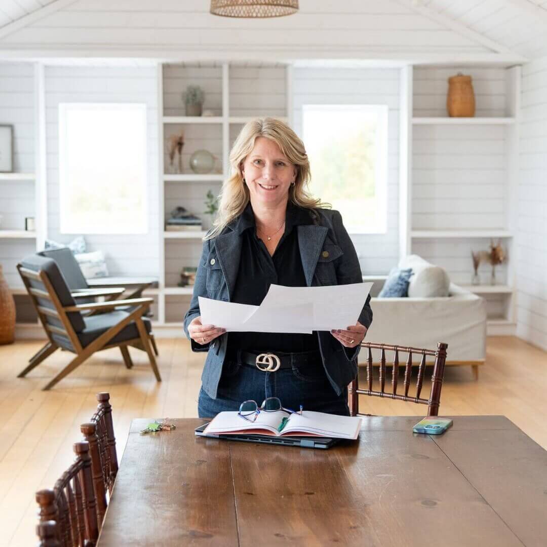 blond femail in a denim jacket standing at a boardroom table holding paper work. The background is a beautiful, white display wall with windows