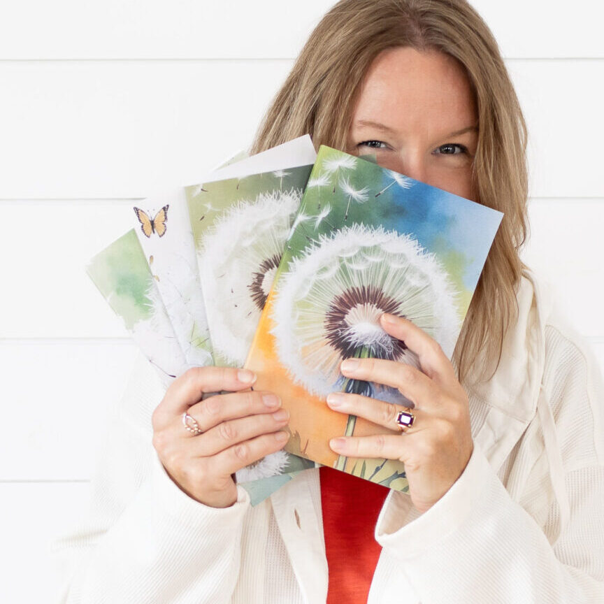 blond women in white jacket over orange tshirt holding up 4 water-colour covered dandelion journals. Journals are a set of 4 Dandelion Drifts by Allison Smith, available on Amazon
